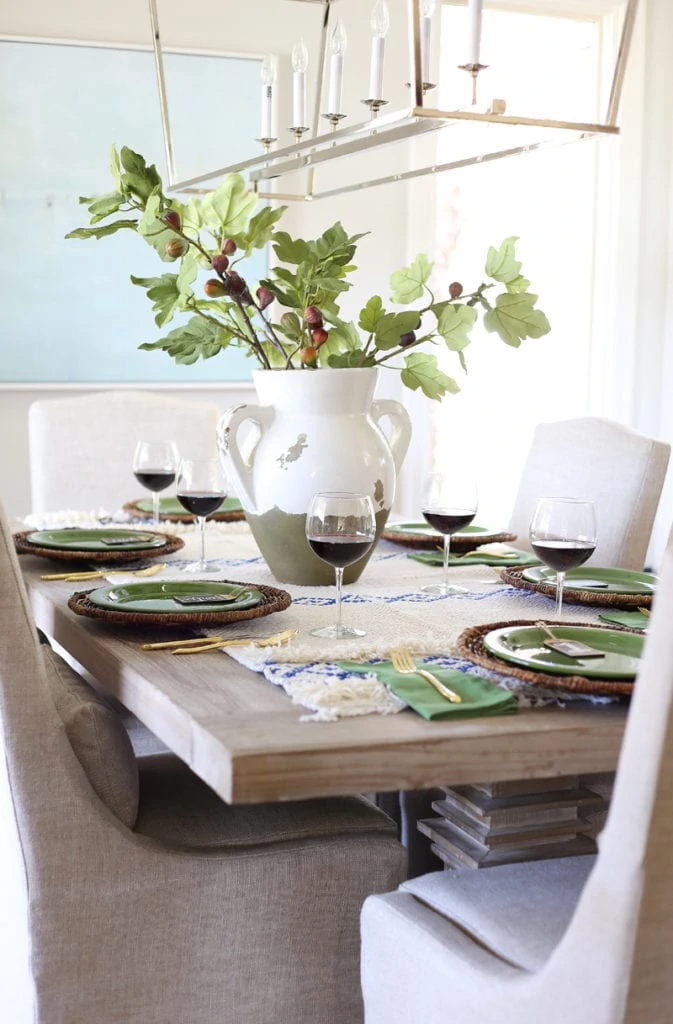 The dining room table with a faux olive branch inside a white and green vase with red wine on the table in glasses.