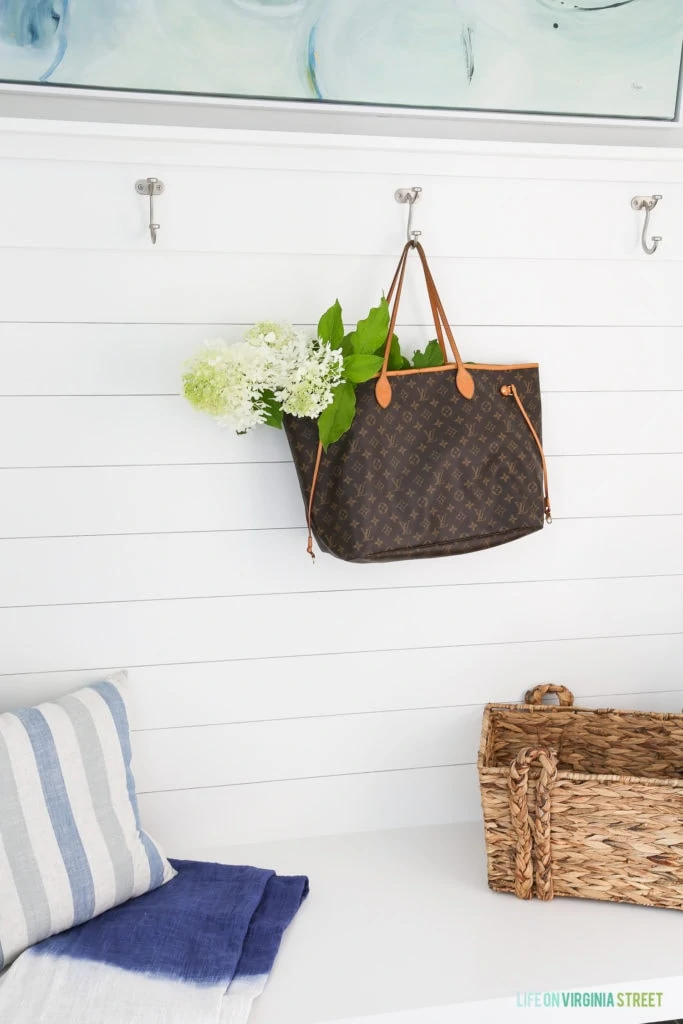 A braided basket is on the bench and a tote bag with flowers hangs on the hooks.