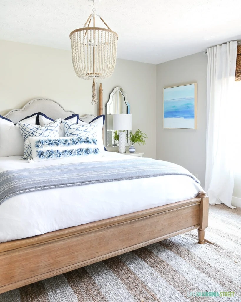 A gorgeous coastal inspired guest bedroom with ocean art, a white bead chandelier, striped rug, and blue and white accents. Such a beautiful and serene bedroom!