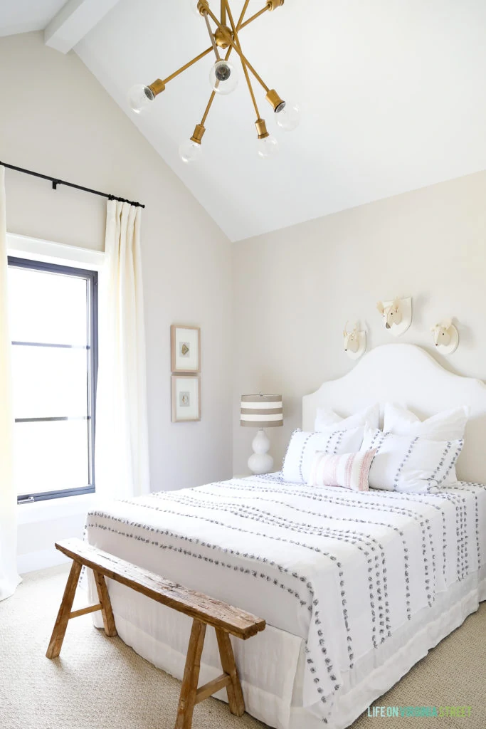 White bedroom with white headboard and white and gray bedding. Gold light fixture and a small wooden bench at the foot of the bed.