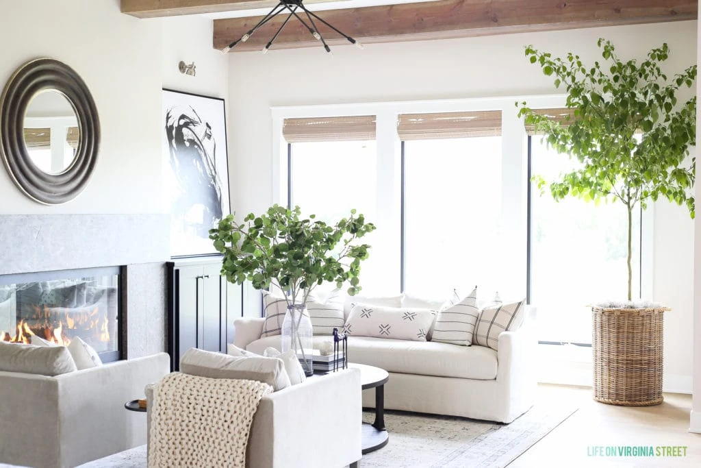A neutral living room with white couches, a mirror on the wall and large green plants in wicker pots.