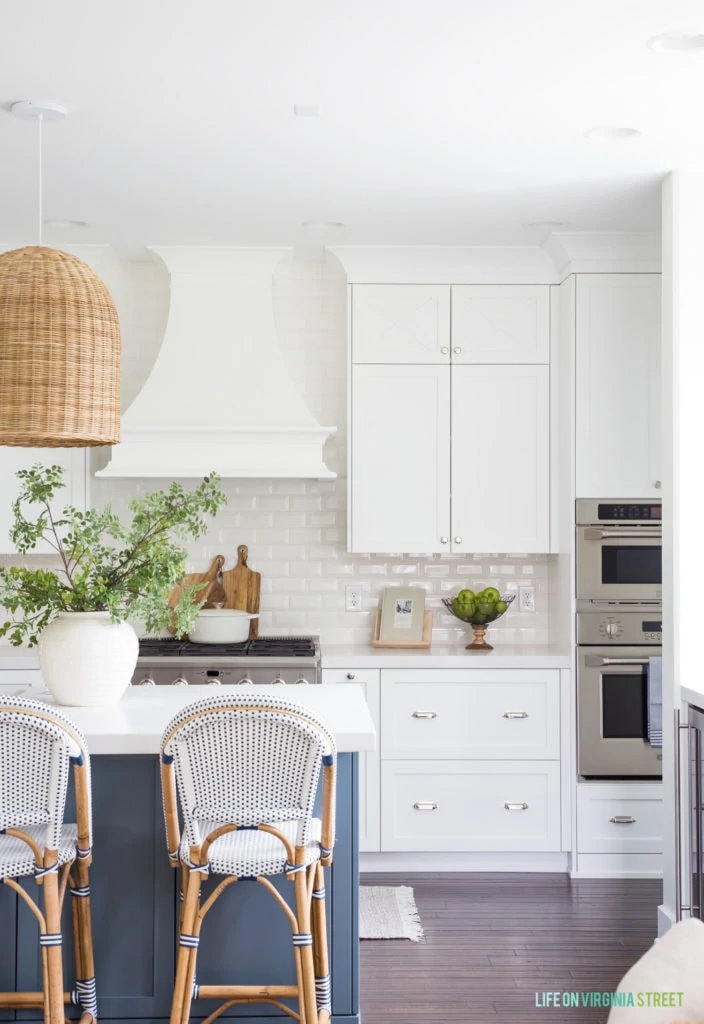 A coastal inspired white kitchen with a blue island. The island has a large white vase with faux greenery underneath a basket pendant light fixture.