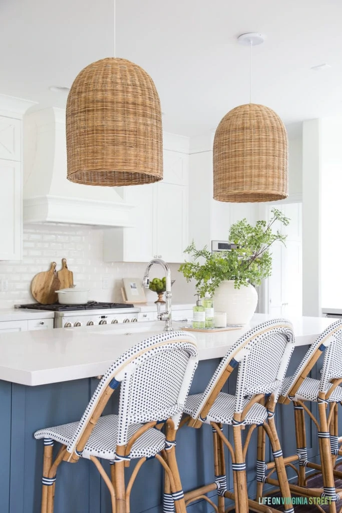 A coastal kitchen with beachy accents including basket pendant lights, a blue island, and woven bistro chairs.