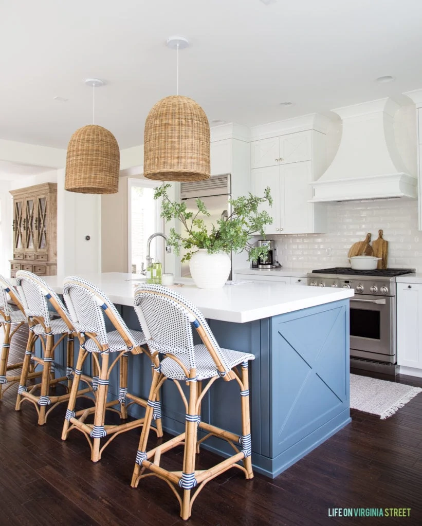 A coastal inspired kitchen with white cabinets, white beveled subway tile, blue island, woven basket pendant lights, blue and white bistro chairs and green and wood toned accents.