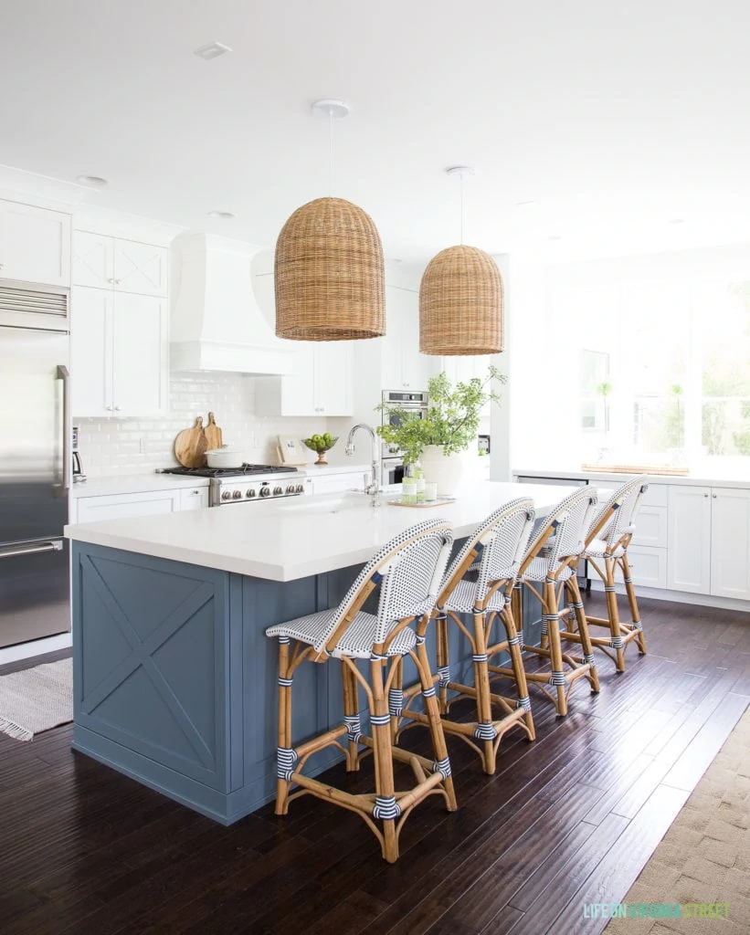 Coastal kitchen with white cabinets, blue island, basket pendant lights, bistro counter stools and a large window. Wall and cabinet colors are Benjamin Moore Simply White, island is Benjamin Moore Providence Blue and countertops are Caesarstone Calacatta Nuvo.