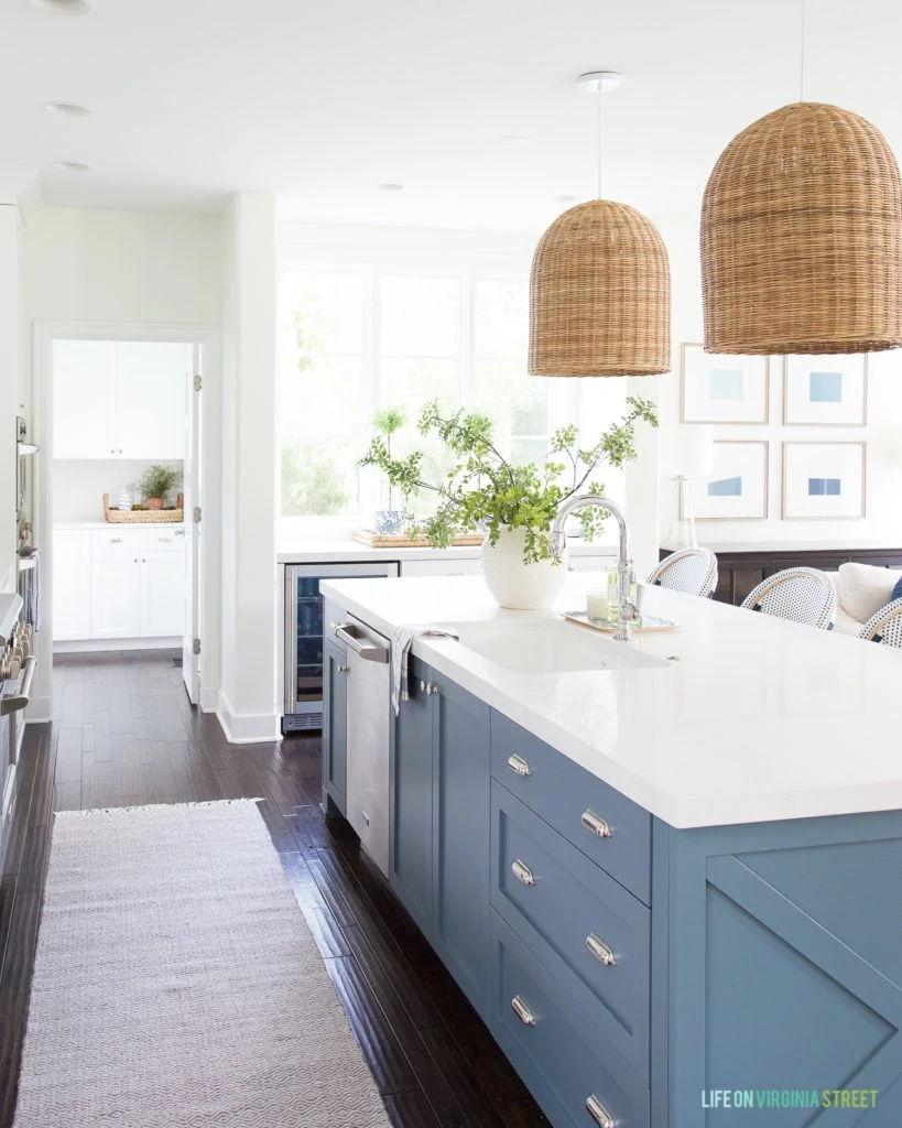 A blue kitchen island with chrome hardware, Caesarstone Calacatta Nuvo countertops, basket pendant lights and green and blue accents.