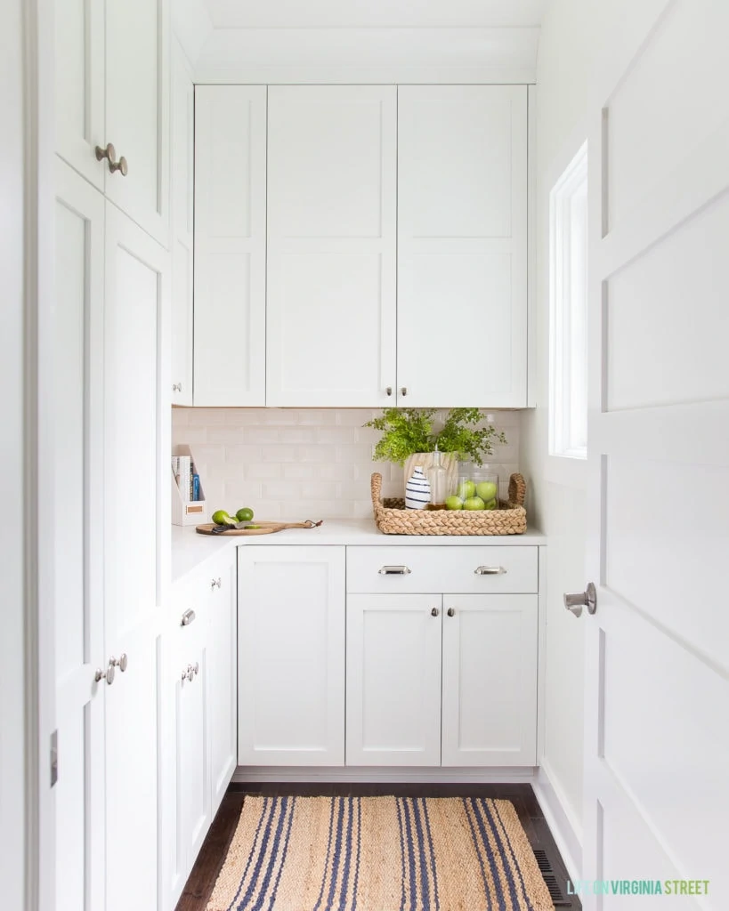 A gorgeous pantry with white cabinets, jute and sisal accents, and a natural and blue striped rug.