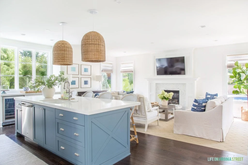 A coastal inspired blue kitchen island and white living room. I love the blue shibori pillows, white spindle chairs, natural woven pendant lights and touches of green.