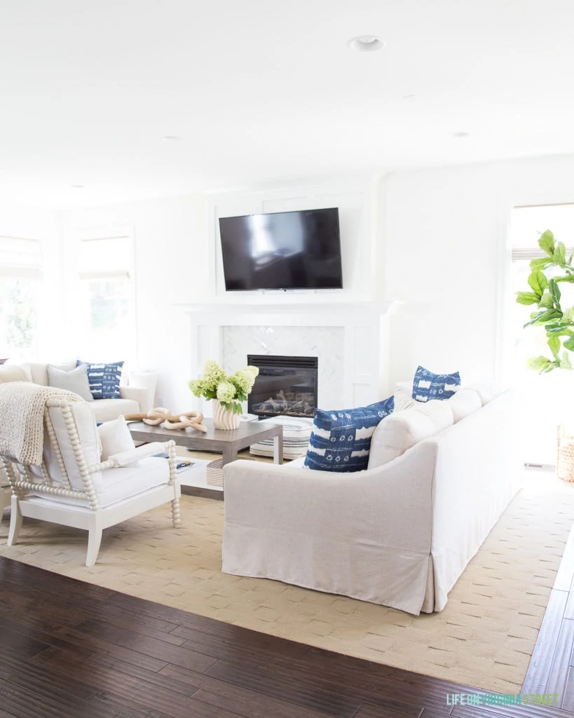 All white living room, which blue and white pillows and a white fireplace with a TV hanging above it.