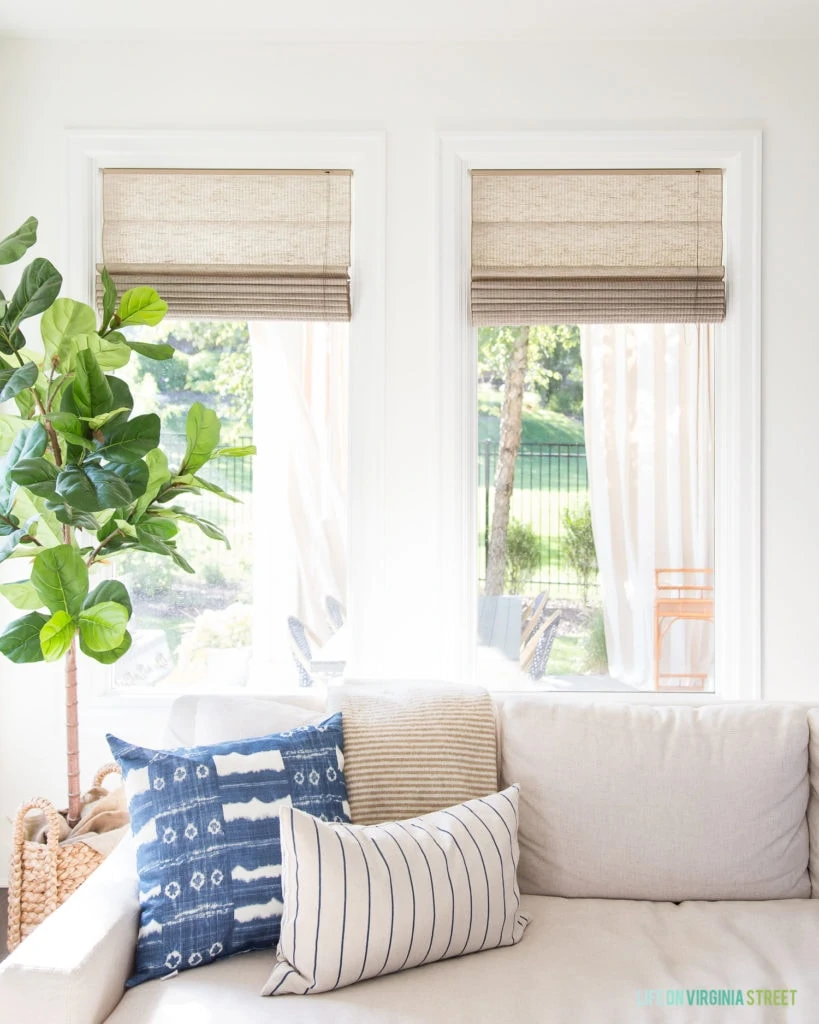 Natural woven roman shades in a living room with blue, white and green accents. Faux fiddle leaf fig tree in corner of the room.