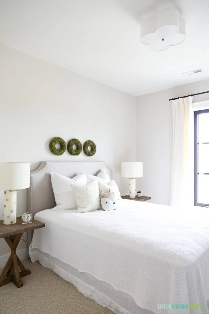A white bedroom with wooden side table and white lamps on them. Three green wreaths above the bed.