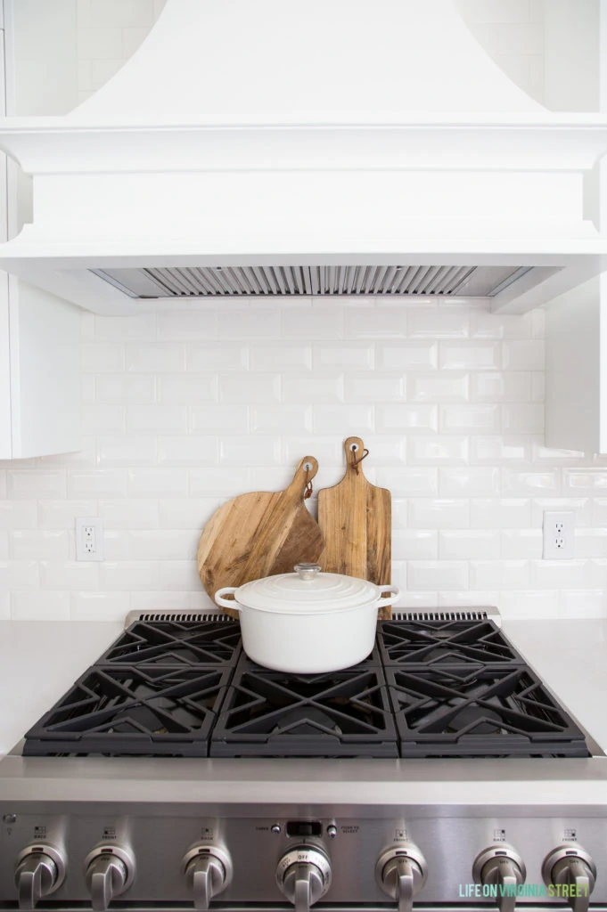 The kitchen stove with a white pot on top of it, wooden cutting boards behind it and white tile on the wall.