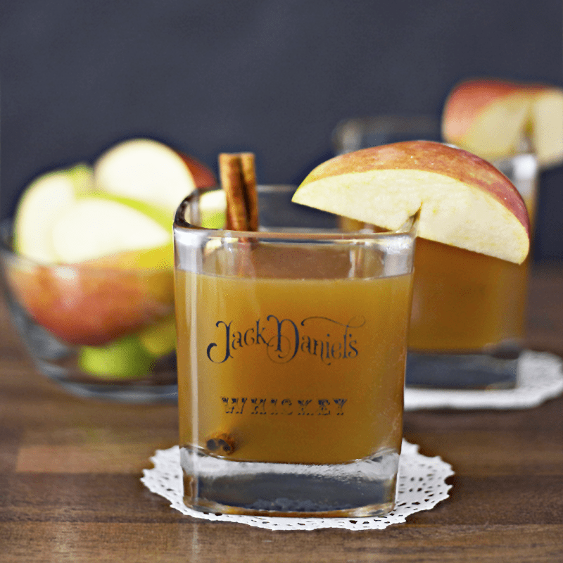 A Jack Daniels whiskey glass cocktail with an apple slice on the rim and cinnamon stick inside.