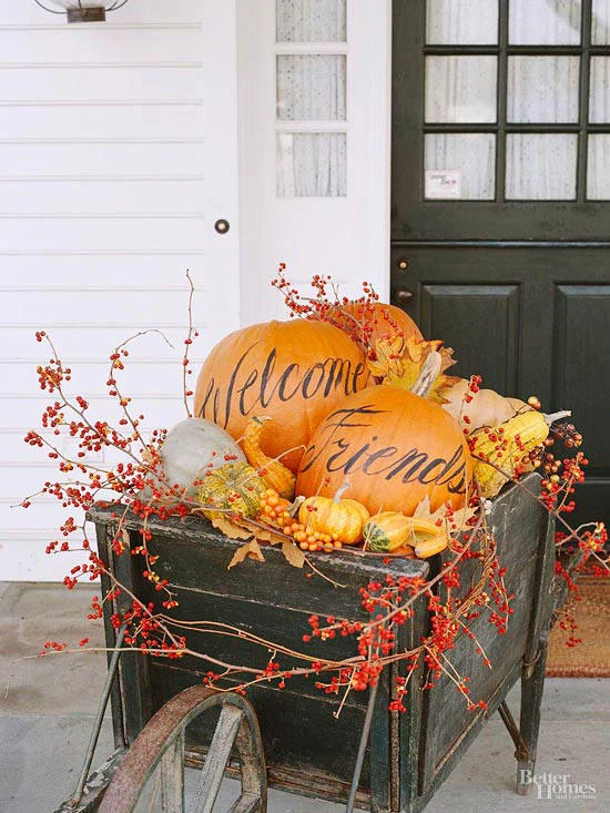 A wooden barrel on the porch filled with pumpkins that have black writing saying Welcome Friends.