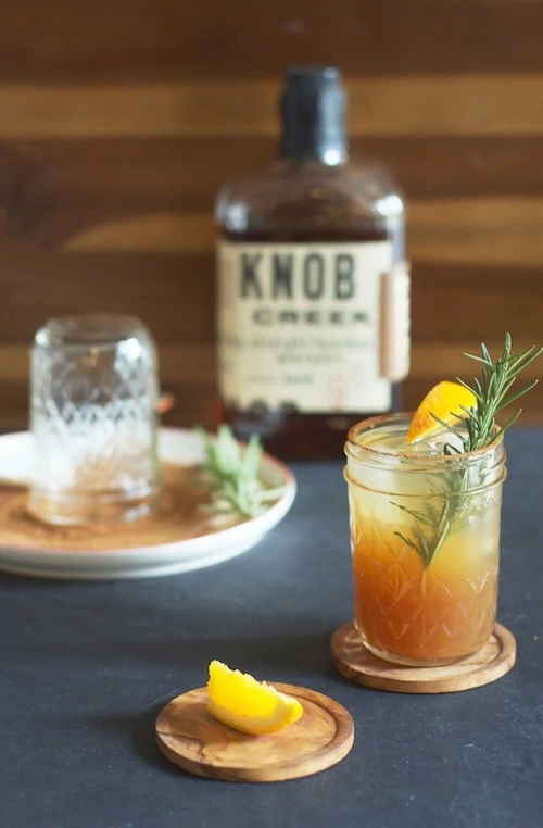 Bourbon drink in a mason jar glass with a sprig of greenery in it and a Knob bourbon bottle in the background.