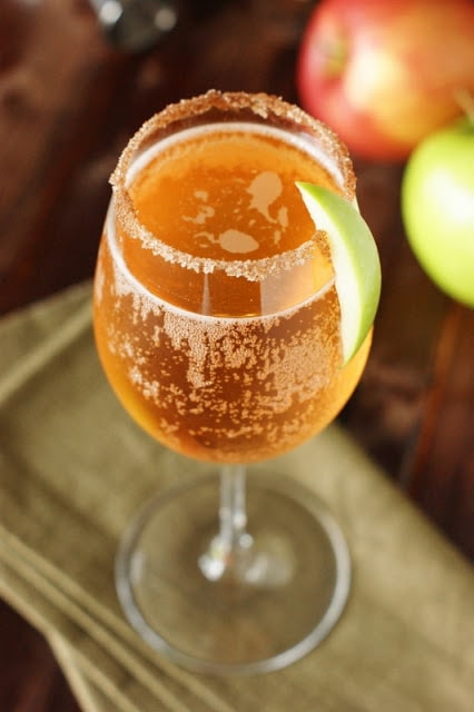 A sparkling cocktail with an apple slice on the rim and sugar on the rim.
