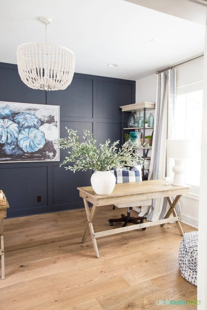 A beautiful home office with navy blue and white walls with white oak hardwood floors. The room is ready for fall with blue pumpkin abstract art, wood desks, a whitewashed wood bead chandelier, and a white vase full of olive leaves.