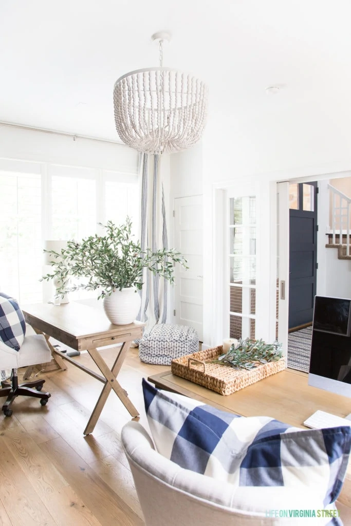 A his and hers home office with white walls, white oak hardwood floors, wood desks, a white wood bead chandelier, faux olive stems, and navy blue buffalo check pillows.