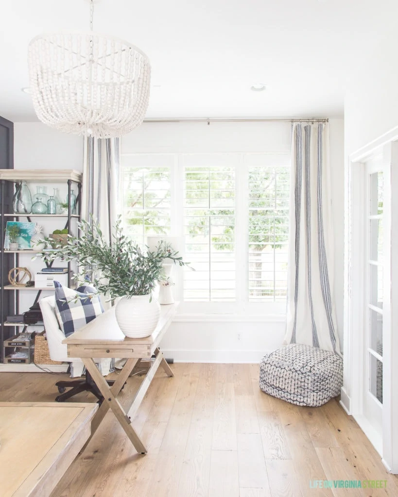 Home office with Benjamin Moore Simply White walls, white oak hardwood floors, blue and white striped curtains, a whitewashed wood bead chandelier, wood desks, navy blue buffalo check pillows and faux olive stems.