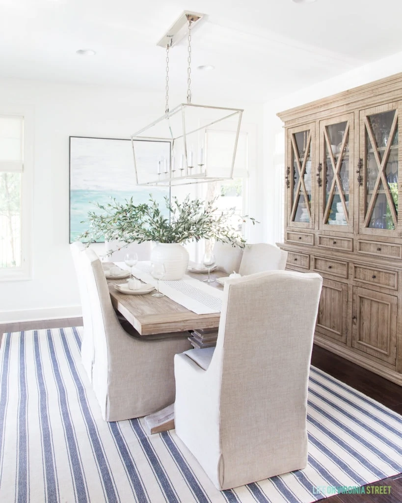 A coastal dining room with a striped rug, linen chairs, reclaimed wood hutch, blue abstract artwork, a Darlana linear pendant chandelier and faux olive leaf stems.