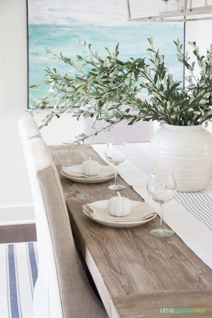 A simply fall tablescape with linen chair, reclaimed wood table, white vase, olive leaf stems, and neutral striped napkins.