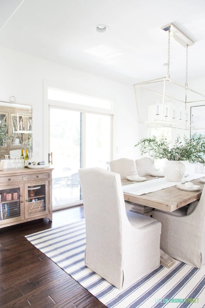 A coastal dining room with blue and white striped rug, Darlana linear chandelier, linen chairs, reclaimed wood dining table and buffet table, a silver mirror, and faux olive leaf stems.