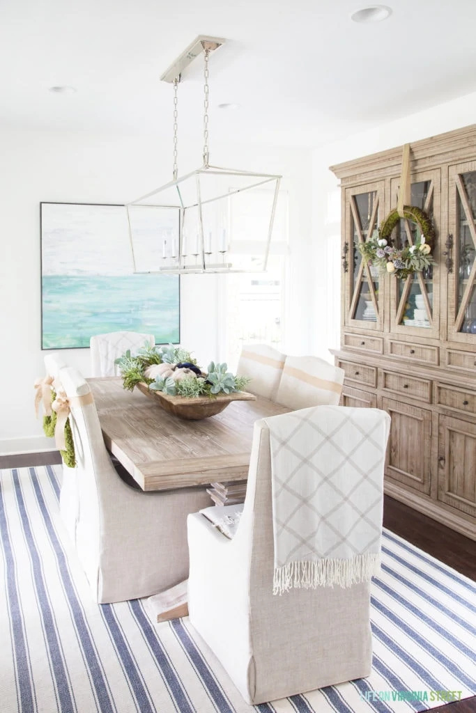 Neutral dining room with a striped rug, white chairs with a wooden table.