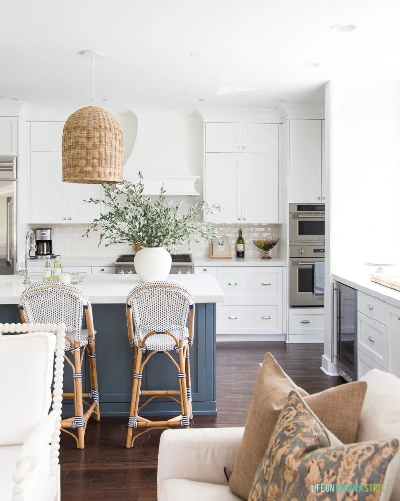 Gorgeous kitchen with white and rich blue cabinets. Love the basket pendant light fixture and the bistro style barstools. The huge pot of olive leaves is perfect for fall!