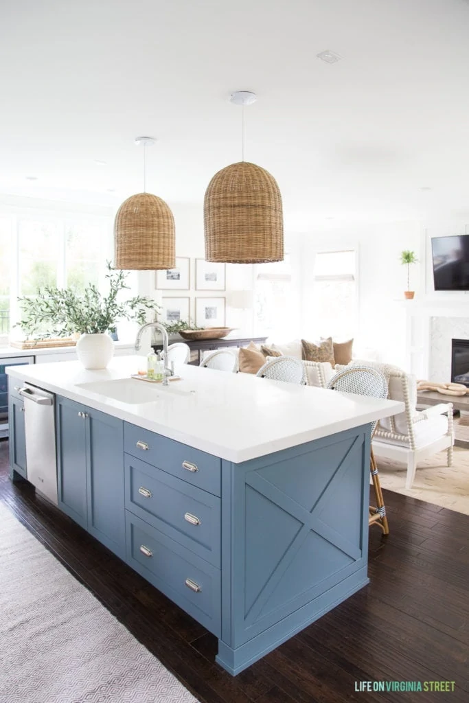 A kitchen decorated for fall. Room has white kitchen cabinets, a blue painted island, basket pendant lights, dark hardwood floors, white walls and a vase filled with olive branches.