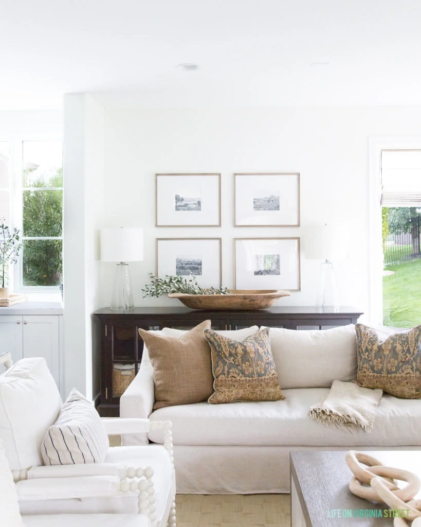 A fall living room with linen sofas, white spindle chairs, a gallery wall with black and white images, and brown and blue accents.