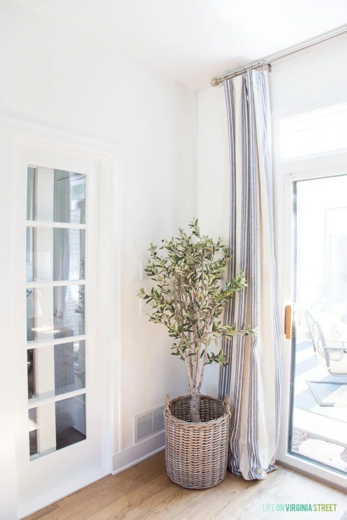 A faux olive tree in a woven basket beside white and blue striped curtains.
