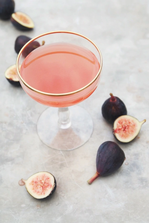 Pink cocktail in a glass with a gold rim and figs lying around it.