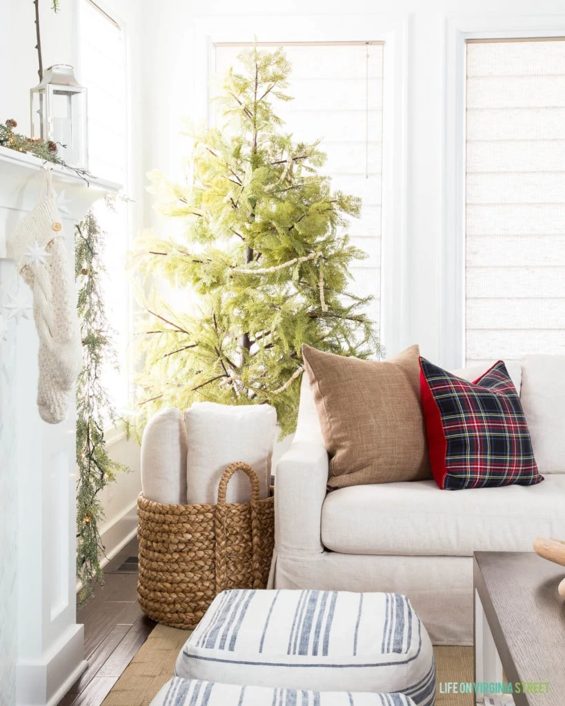 A linen sofa with brown linen pillow, black Stewart plaid pillow, a faux Cypress tree, a woven natural basket, silver lanterns, and chunky knit stockings are perfect in this Christmas living room!