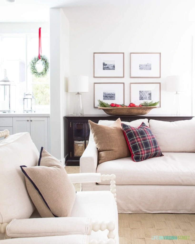 A neutral Christmas living room with white spindle chairs, a linen sofa, brown linen pillow, black and red Stewart plaid pillow, wood gallery wall, wood dough bowl filled with ornaments and greenery, and wreaths hung in the window. Such a cute Christmas family room!