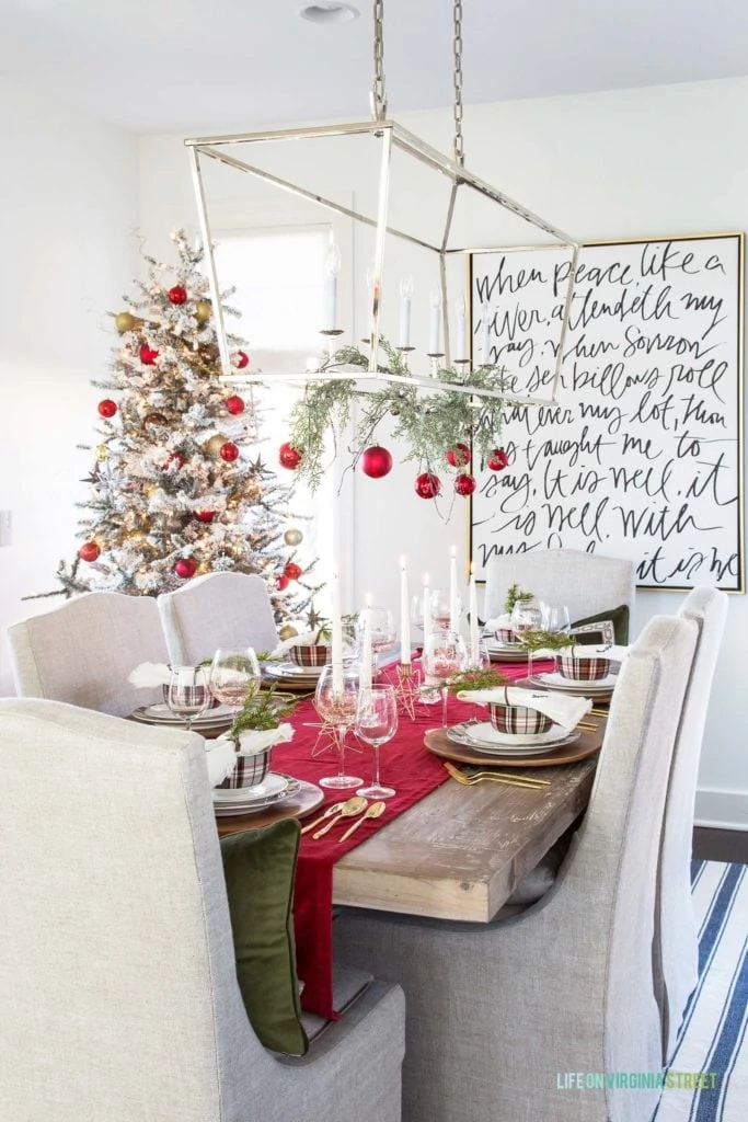 A Christmas tablescape with a red velvet runner, gold flatware and plaid plates and bowls. I love the garland and ornaments hanging from the silver chandelier!