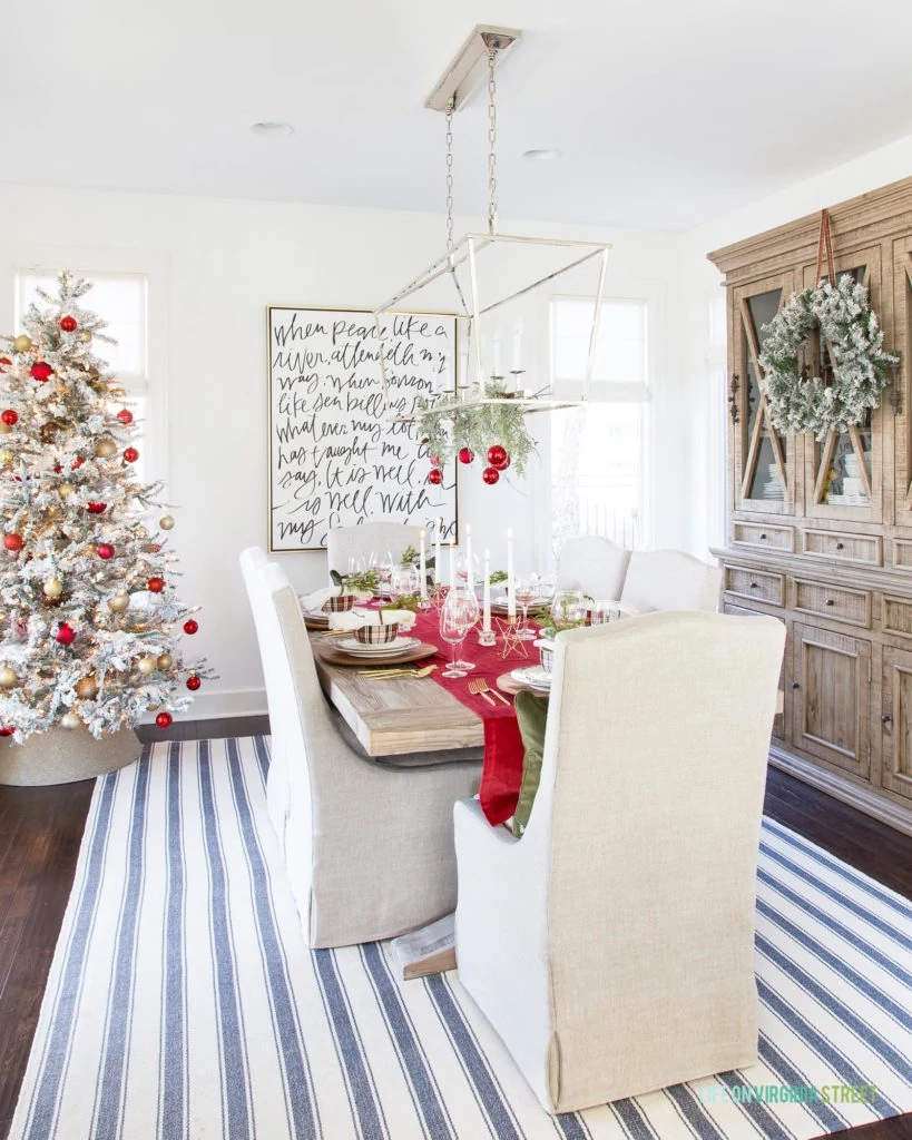 A coastal dining room with linen chairs, reclaimed wood table, silver linear Darlana pendant light decorated with garland and red ornaments, a flocked Christmas tree and a blue and white striped rug. I love the large "It Is Well" canvas art on the wall!