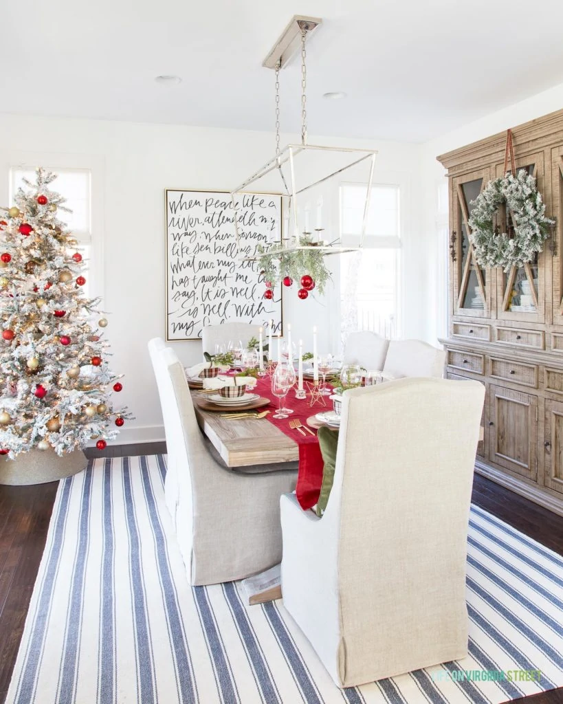 A Christmas dining room with a red plaid Christmas tablescape, a blue and white rug and wooden hutch in the dining room.