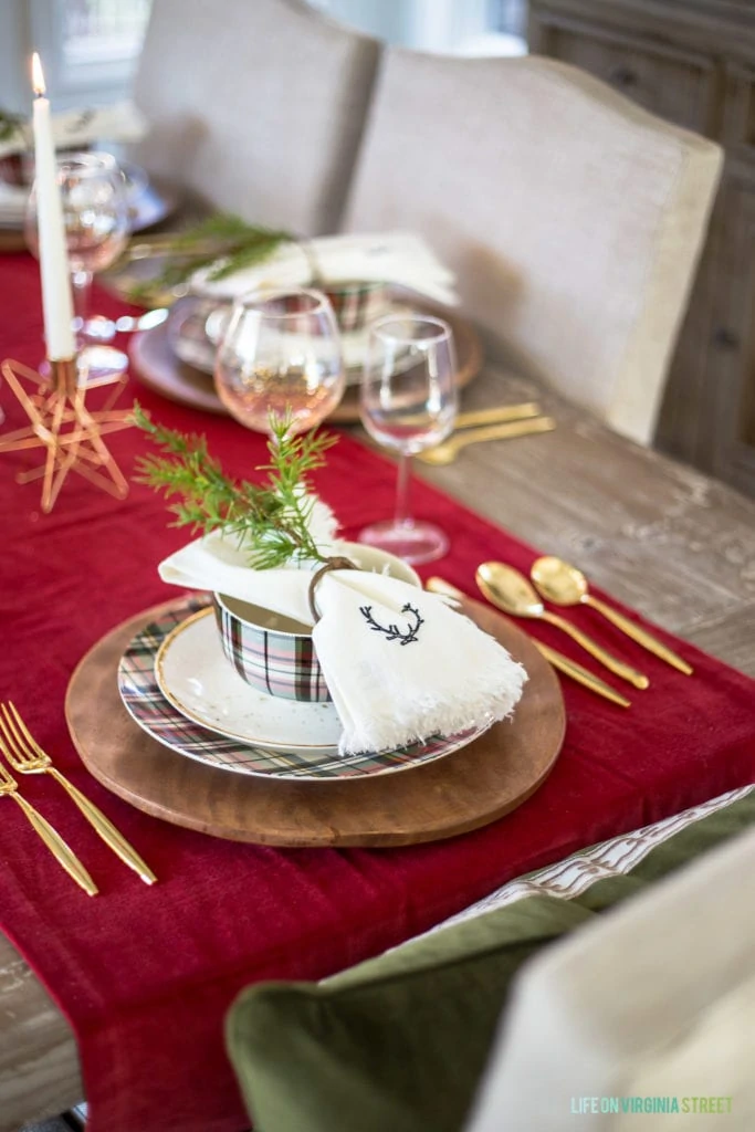 A Christmas place setting with plaid plates, gold flatware, white linen deer napkins, and faux greenery napkin rings.