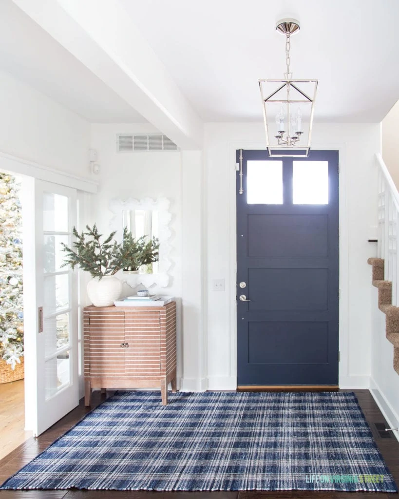 A wintery Christmas entryway with navy blue front door, white walls, a navy blue plaid rug, striped wood cabinet, silver lantern pendant light, and faux greenery. You can also see a peek of the blue and metallic flocked Christmas tree!