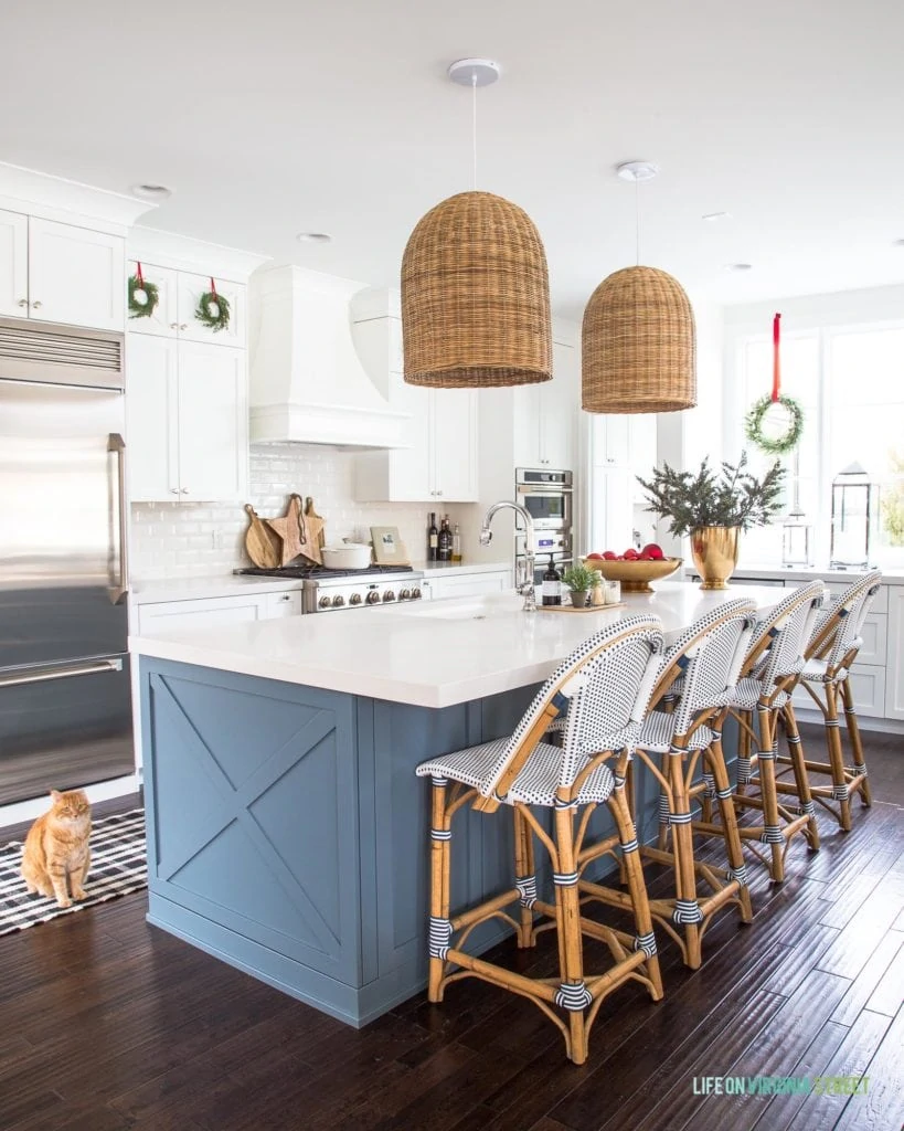 A Christmas kitchen with small wreaths on the cabinets hung with red velvet ribbons. I love the white cabinets with the white beveled subway tile, blue kitchen island, basket pendant lights, and French bistro style counter stools! The gold bowl and gold vase filled with ornaments and greenery add a festive vibe!