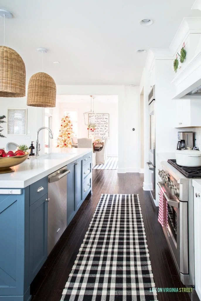 A beautiful Christmas kitchen with white cabinets, a blue kitchen island, a black and white plaid runner, basket pendant lights, and a view of the flocked Christmas tree in the dining room!