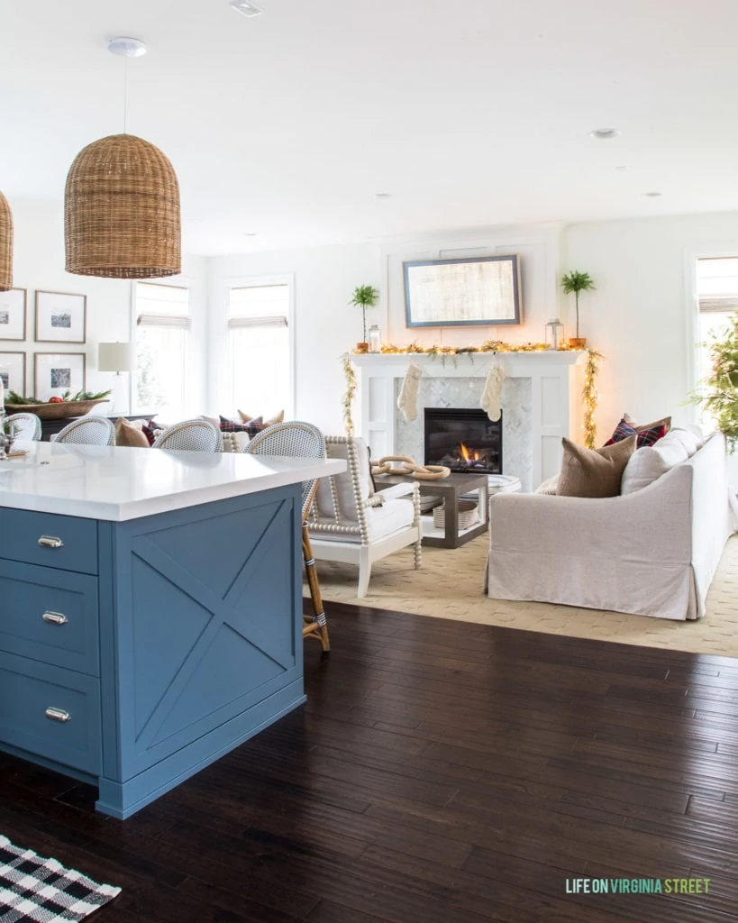 A Christmas kitchen and living room. The kitchen has a blue island, white quart countertops, basket pendant lights and a black and white plaid runner. The living room has linen sofas, and black and red plaid pillows! I love the garland and knit stockings on the white fireplace!