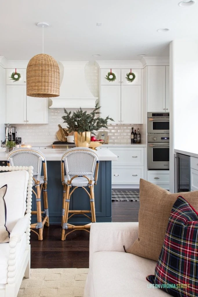A white Christmas kitchen with blue island, wreaths on the cabinet doors, basket pendant lights, plaid pillows, bistro chairs and plaid rugs!