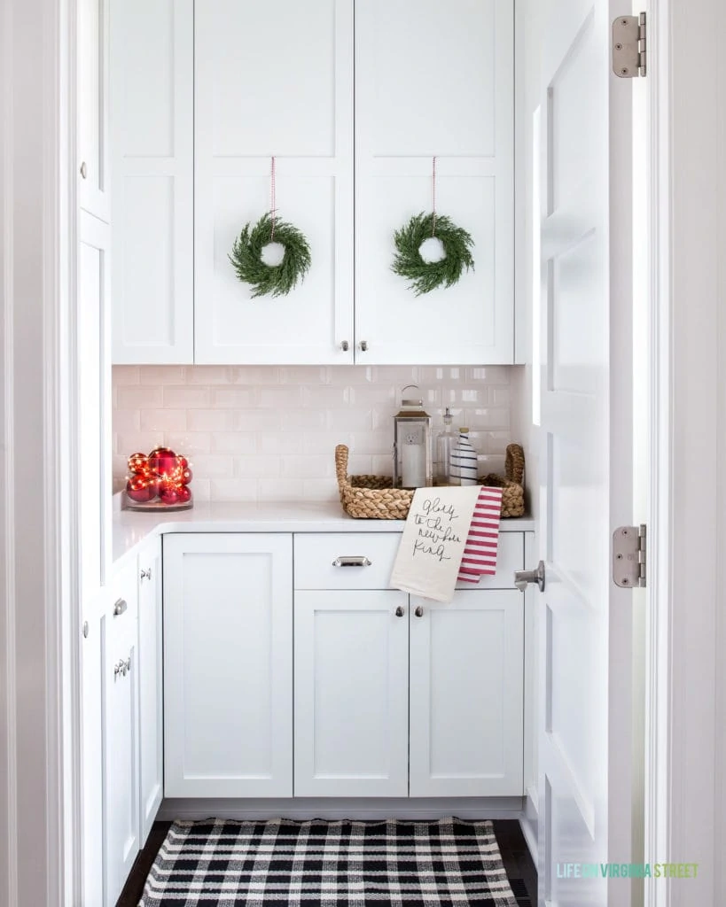 The cutest kitchen Pantry decorated for Christmas! I love the idea of cabinets in a pantry, and these mini green wreaths, black and white plaid rug, red ornaments and other festive touches are the perfect addition!