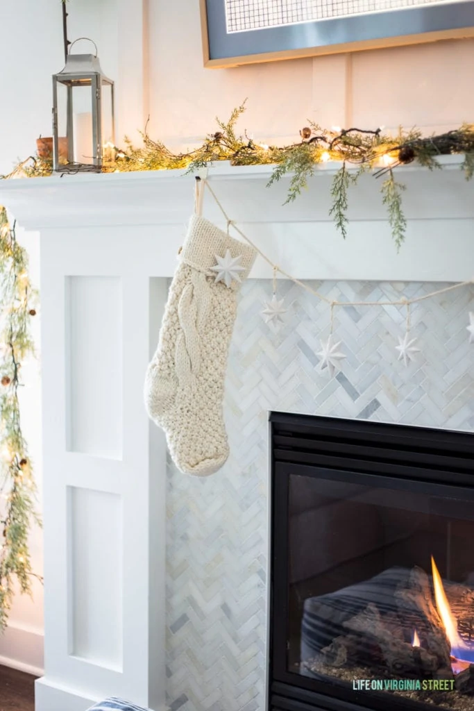A chunky knit stocking hung on a white fireplace mantle with herringbone marble tile surround. I love the garland and lights along with the silver lantern!