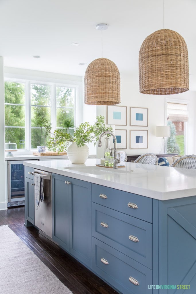 A coastal inspired kitchen with a Benjamin Moore Providence Blue island. Also includes basket pendant lights, bistro style bar stools, and a large window overlooking the greenery outside.