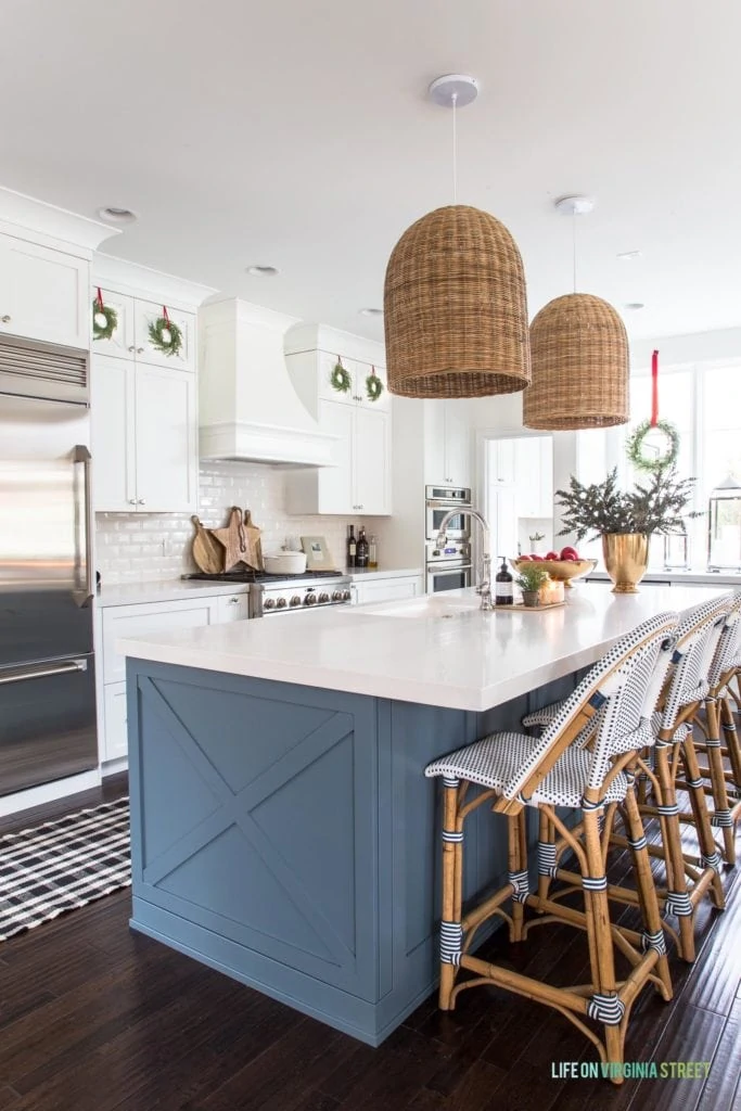 A white kitchen with a blue island, basket pendant lights, mini wreaths on cabinets, gold vase, and black and white runner. Such a cute Christmas kitchen!