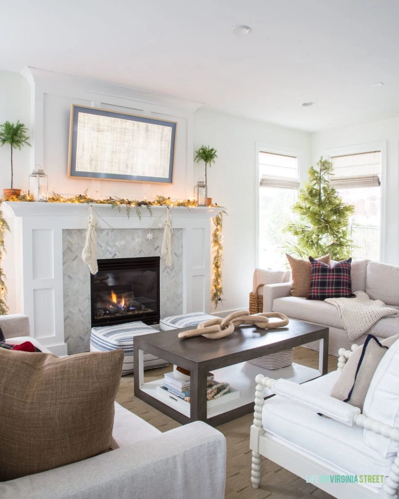 A Christmas living room with neutral linen sofa and chairs, a white mantle, and herringbone marble fireplace surround. The garland with twinkle lights, faux Cypress tree, and black Stewart plaid pillows add a fun festive touch to this family room.