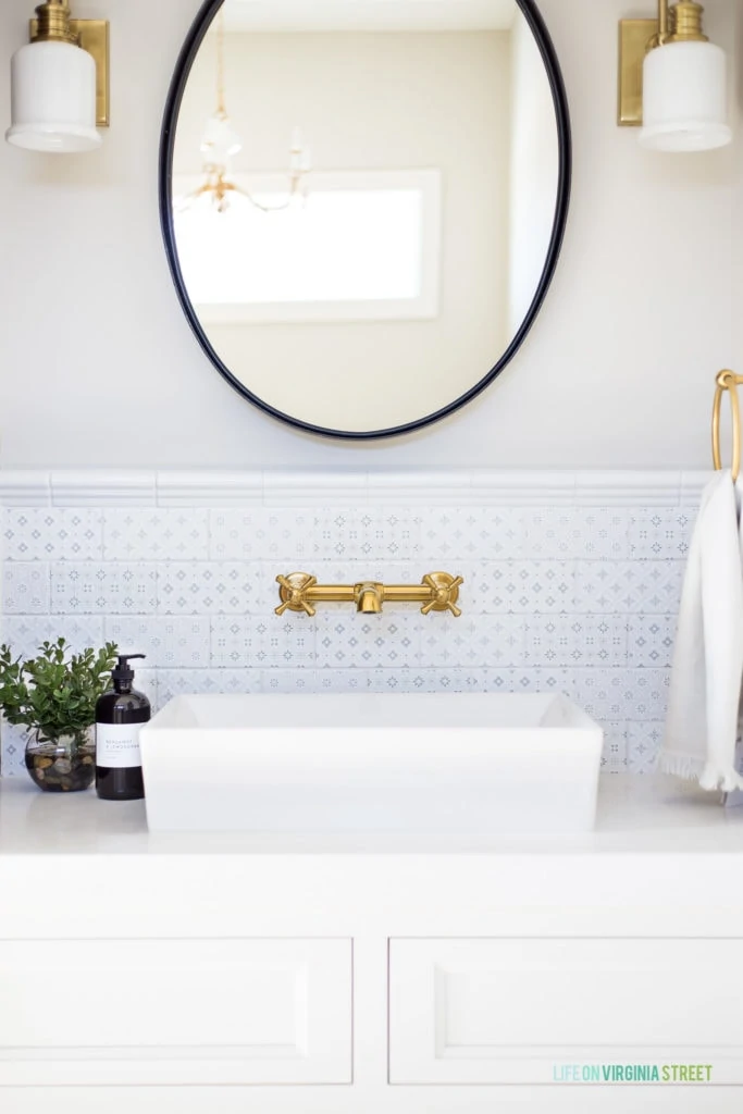 A beautiful vanity with white cabinets, blue and white patterned subway tile, iron mirror, brass sconces and gold fixtures and hardware.
