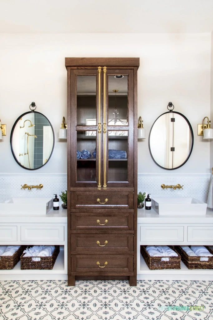 A double white vanity with walnut stained linen cabinet, oval iron mirrors, blue and white patterned backsplash tile, and gold fixtures.